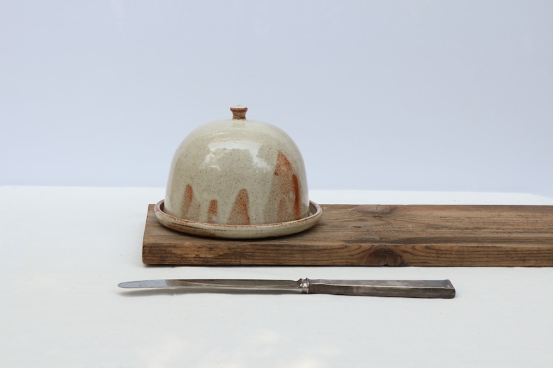 May include: A white ceramic butter dish with a brown glaze and a dome lid. The dish is sitting on a wooden board with a silver butter knife.