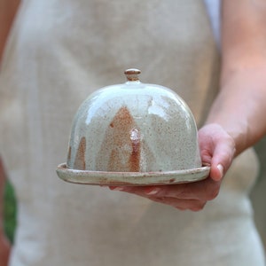 May include: A ceramic butter dish with a dome lid. The dish is glazed in a light gray color with brown speckles. The lid has a small knob on top.