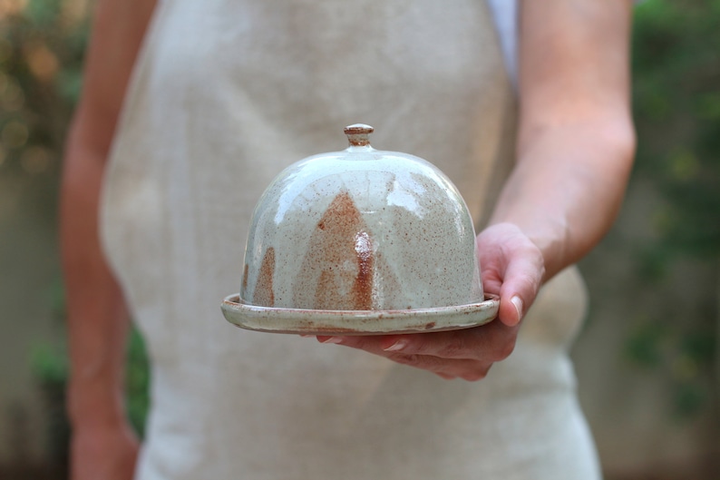 May include: A ceramic butter dish with a dome lid. The dish is glazed in a light gray color with brown speckles. The lid has a small knob on top.
