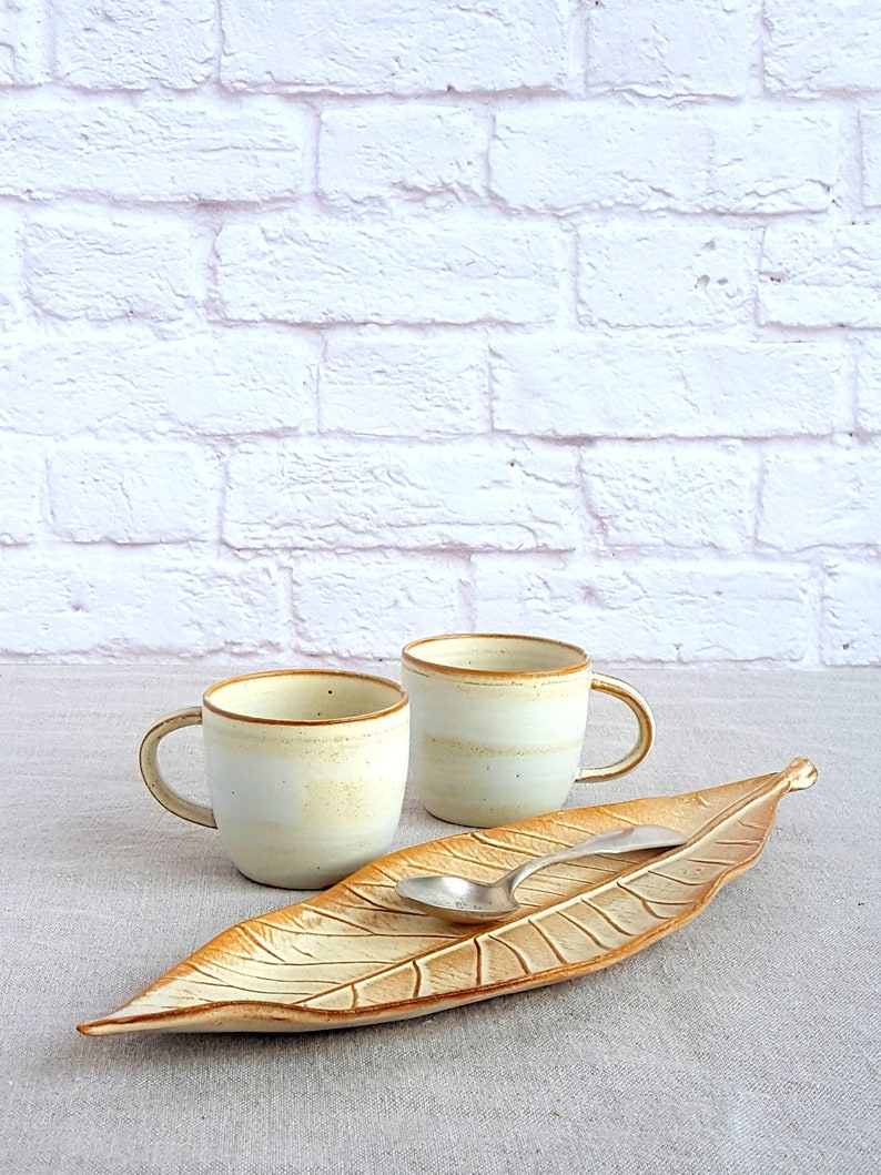 May include: Two white ceramic mugs with a brown rim and a brown leaf-shaped serving dish with a silver spoon.
