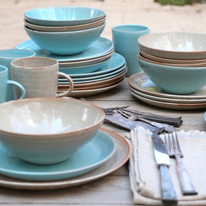 Puede incluir: Un conjunto de vajilla de cerámica en tonos azules y beige, que incluye cuencos, platos y tazas. La vajilla de estilo rústico tiene un acabado texturizado y un borde marrón. También hay cubiertos y una servilleta de lino.