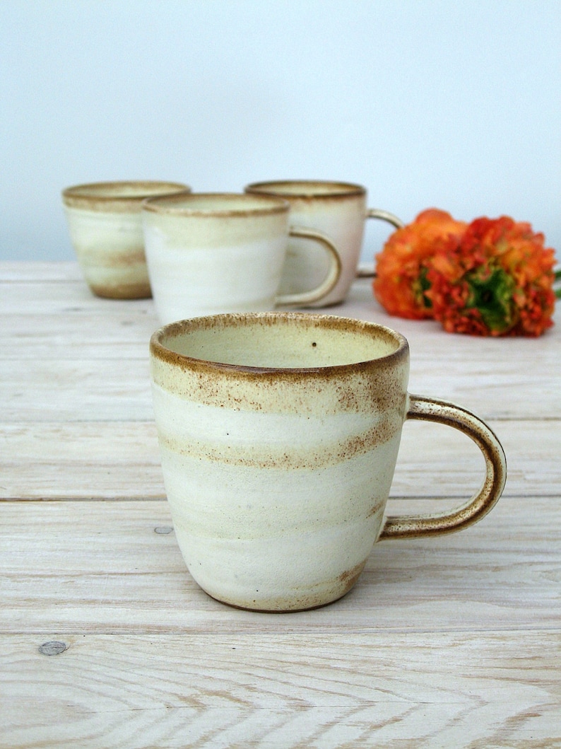 May include: Four ceramic mugs with a white glaze and brown speckles. The mugs have a rounded shape and a handle. The mugs are arranged in a row on a light wood surface.