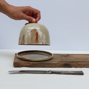 May include: A ceramic butter dish with a lid, the lid is lifted and held by a hand. The dish is on a wooden board with a silver butter knife.
