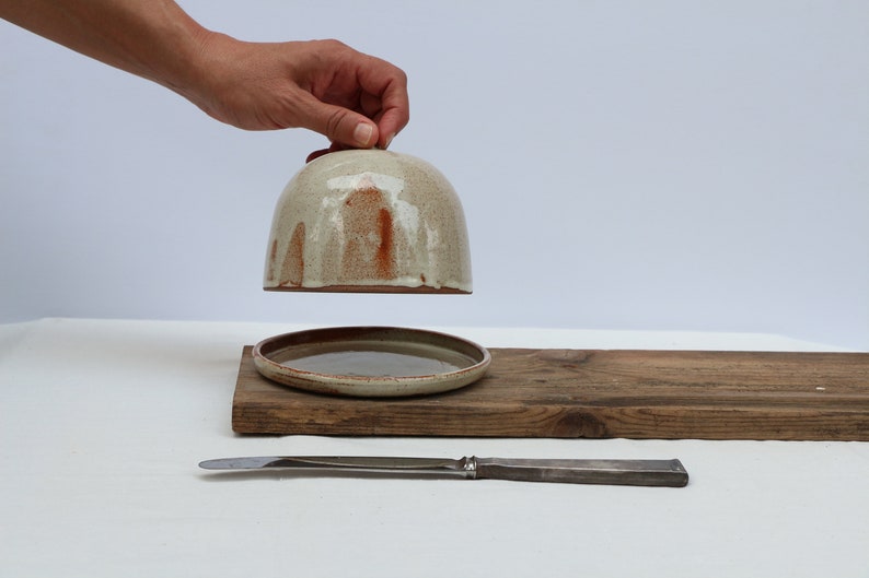 May include: A ceramic butter dish with a lid, the lid is lifted and held by a hand. The dish is on a wooden board with a silver butter knife.