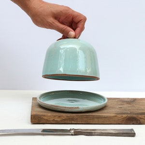 May include: A light blue ceramic butter dish with a lid and a matching plate. The dish is sitting on a wooden board with a silver butter knife.