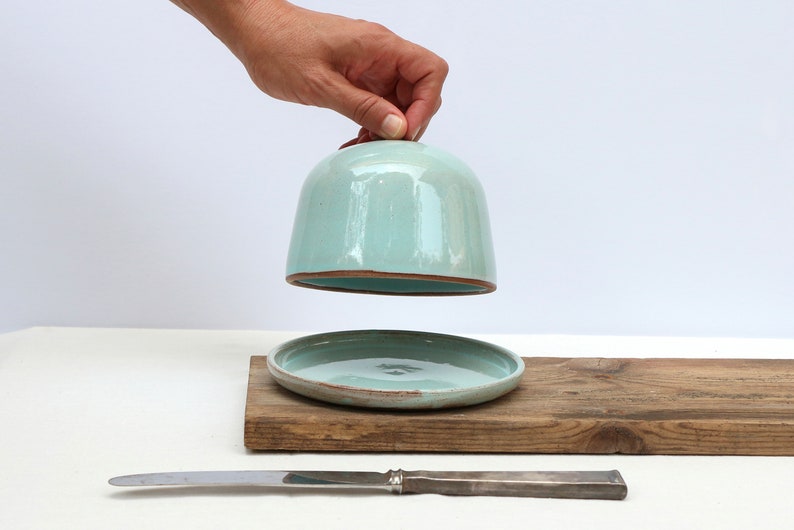 May include: A light blue ceramic butter dish with a lid and a matching plate. The dish is sitting on a wooden board with a silver butter knife.