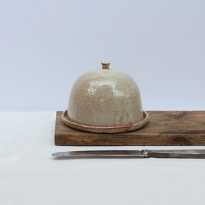 May include: A ceramic butter dish with a dome lid. The dish is a speckled beige color with a brown rim. The dish is sitting on a wooden board.