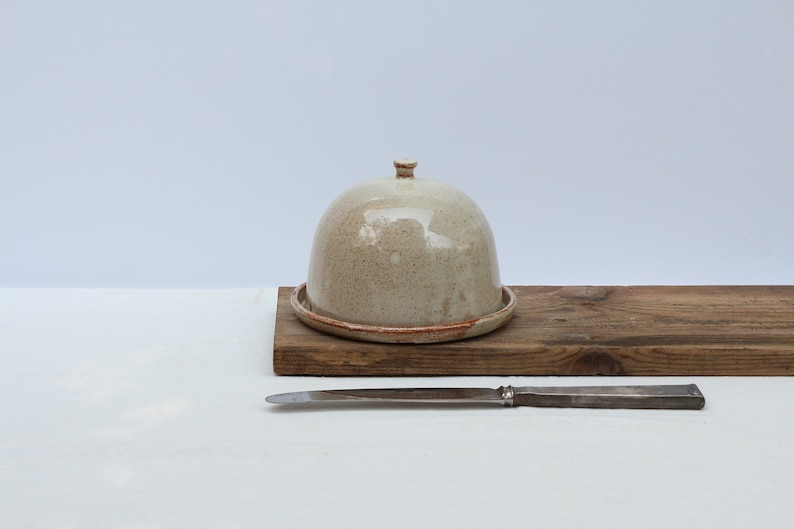 May include: A ceramic butter dish with a dome lid. The dish is a speckled beige color with a brown rim. The dish is sitting on a wooden board.