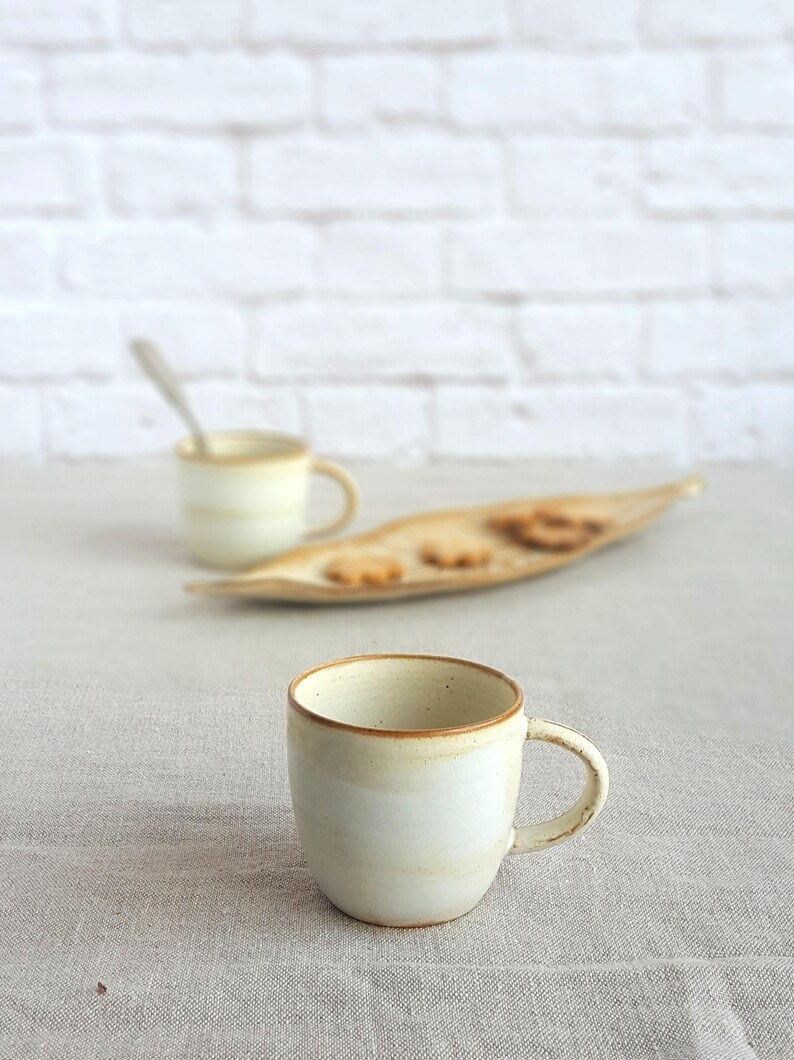 May include: Two white ceramic mugs with brown rims. The mug in the foreground is on a light gray linen tablecloth. The mug in the background is out of focus and is on a wooden tray with cookies.