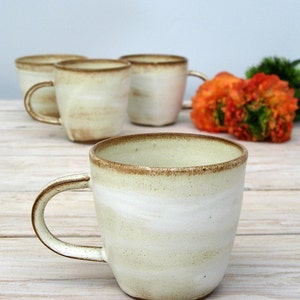 May include: Four ceramic mugs with a white glaze and brown rim. The mugs have a speckled, marbled effect. The mugs are arranged in a row on a light wood surface.