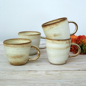 May include: Four ceramic mugs with a white glaze and brown speckles. The mugs have rounded handles and are stacked on a light wood surface.