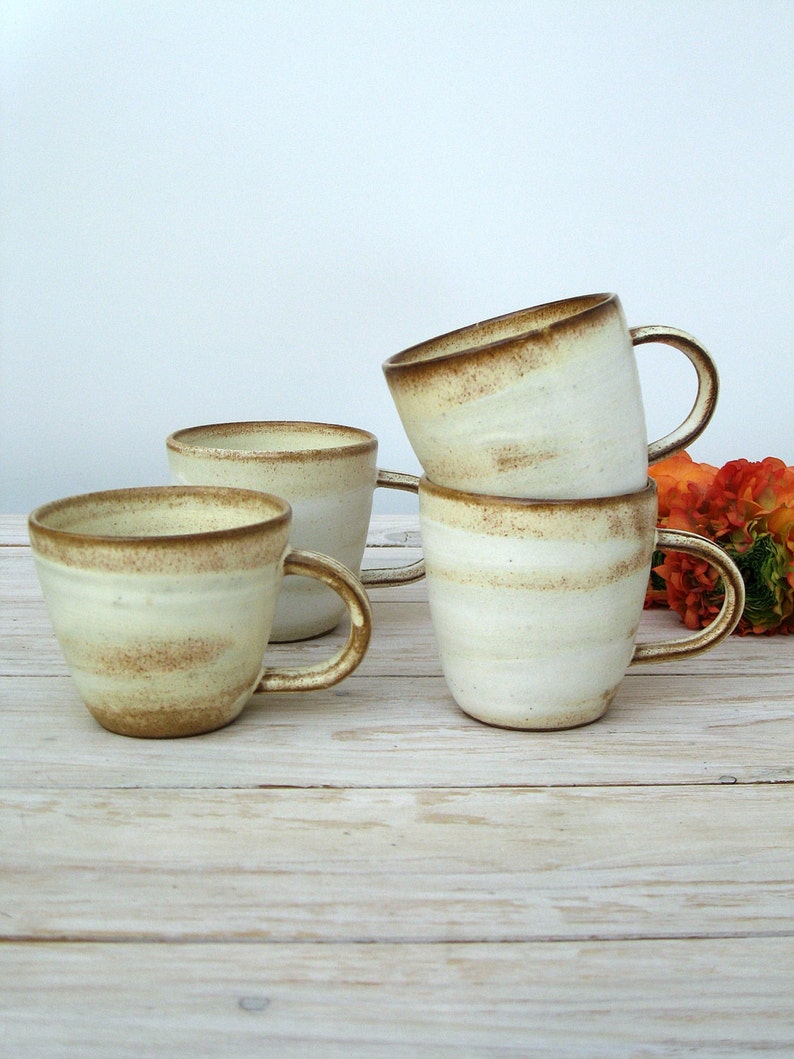 May include: Four ceramic mugs with a white glaze and brown speckles. The mugs have rounded handles and are stacked on a light wood surface.
