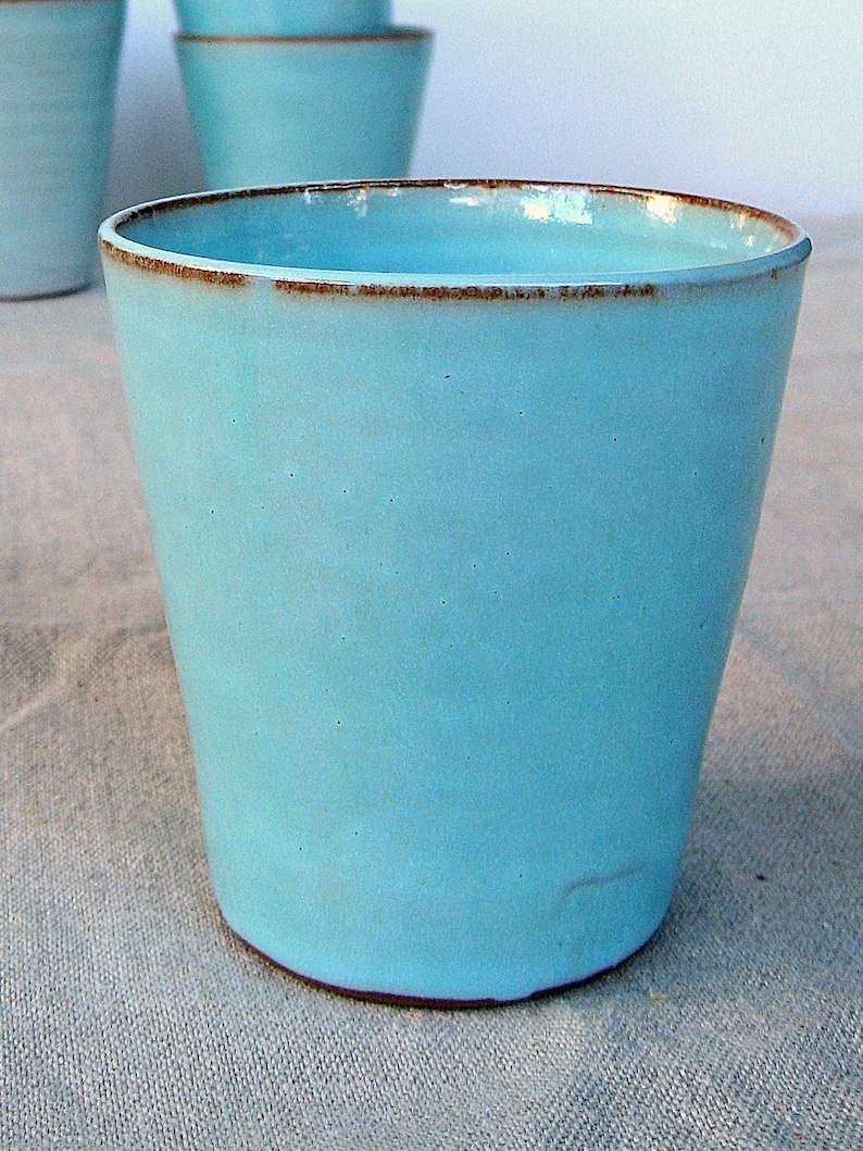 May include: A light blue ceramic mug with a brown rim. The mug is sitting on a light brown fabric surface.