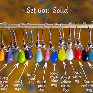 Könnte beinhalten: Set mit 12 Weinglasanhängern mit verschiedenen farbigen Glas Tropfen. Die Anhänger sind mit Nummern und Farbnamen beschriftet, wie z. B. "601-1 orange #50", "601-2 lila Streifen #40" und "601-3 grüner Streifen #62".