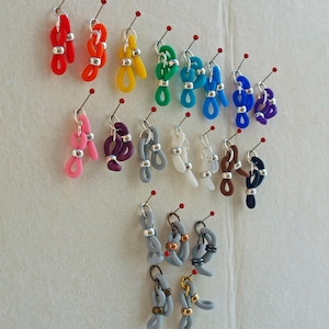 May include: Assortment of colorful silicone glasses strap holders. The holders are in various colors, including red, orange, yellow, green, blue, purple, pink, gray, white, brown, and black. The holders are attached to silver pins.