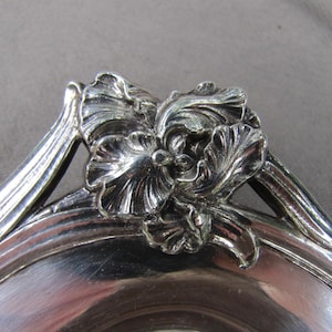 May include: A polished silver-toned metal dish featuring a prominent three-dimensional floral relief ornament on the rim. The flower design displays intricate, layered petals with deep grooves and a reflective, metallic finish throughout the piece.