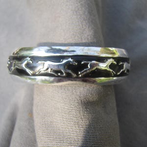 May include: Silver cuff bracelet with a black enamel band featuring a repeating pattern of running horse silhouettes. The bracelet has a polished finish and a classic design. The bracelet is approximately 2.5 cm wide.