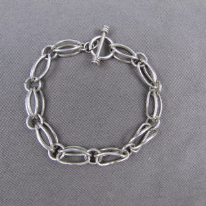 May include: A silver-toned metal chain bracelet featuring a series of interconnected oval links. The bracelet is secured with a toggle clasp consisting of a circular ring and a perpendicular bar, creating a continuous circular design.