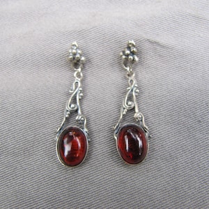 May include: A pair of silver-toned dangle earrings. Each earring features an oval, deep red gemstone set in a decorative frame. The earrings have a floral design at the top and a swirling pattern connecting to the gemstone.