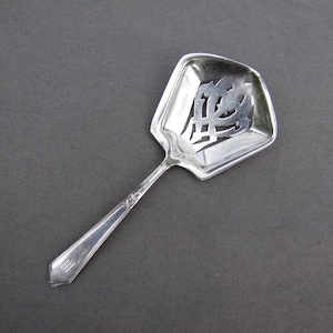 May include: A silver-colored serving spoon with a decorative openwork design in the bowl. The spoon has a long, slender handle with a subtle embossed pattern near the bowl. The bowl is square with a beveled edge.