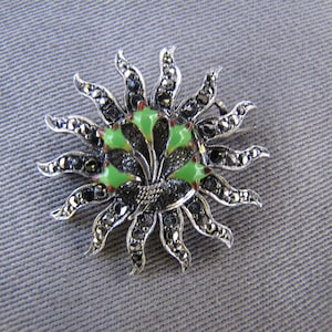 May include: A silver-toned sunburst brooch with a central design of green enamel and black accents. The brooch features radiating rays adorned with small, dark stones, creating a striking contrast against the gray background.