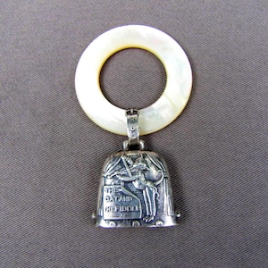 May include: A silver rattle featuring a bell with an embossed illustration of a cat playing a fiddle and the text THE CAT AND THE FIDDLE. The bell is attached to a smooth, circular, off-white mother-of-pearl teething ring by a silver loop.