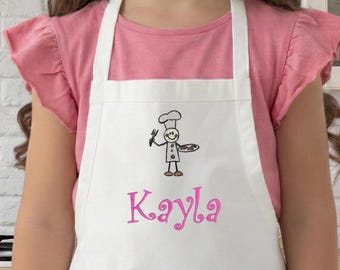May include: A young person wearing a white apron with a chef graphic and the name "Kayla" in pink script. The apron is worn over a pink t-shirt. The background includes a kitchen setting with a white brick wall and a range hood.