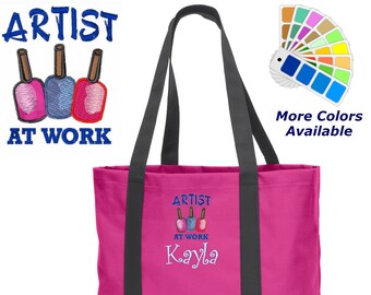 May include: Pink tote bag with black straps and a bottom. The bag has an embroidered design of three nail polish bottles with the text "Artist at Work Kayla".