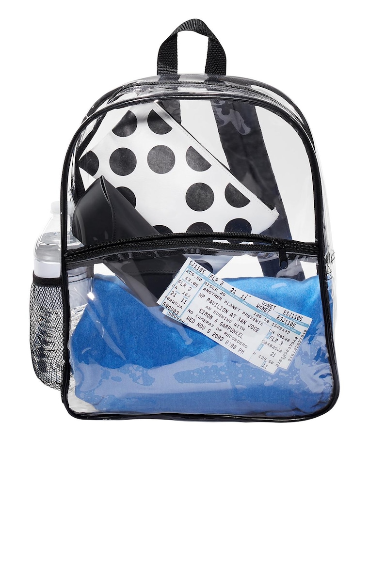 May include: Clear plastic backpack with black trim and zipper pulls. The backpack is filled with various items, including a black and white polka dot pouch, a blue blanket, a water bottle, and a concert ticket for Simon & Garfunkel. The ticket reads "WED NOV 5 2003 8:00 PM".