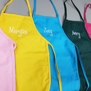 May include: A row of colorful aprons in pink, yellow, blue, green, and fuchsia. Each apron has a white embroidered name: Morgan, Zoey, Avery, and Evelyn. The aprons have neck straps and side ties.