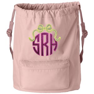 May include: A dusty rose drawstring backpack featuring a front pocket and adjustable shoulder straps. The center displays an embroidered monogram with the letters SRH in dark purple glitter, topped with a lime green and silver glitter bow.