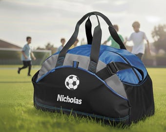 May include: A blue, black, and gray duffel bag featuring a white soccer ball graphic and the name Nicholas printed in white text. The bag includes black carrying handles, a side mesh pocket, and blue accent stitching on the exterior fabric.