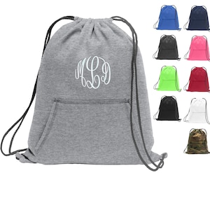 May include: A gray drawstring bag with a large white monogram on the front. The bag has a pocket on the front and black drawstrings. The bag is shown against a white background with other drawstring bags in various colors.