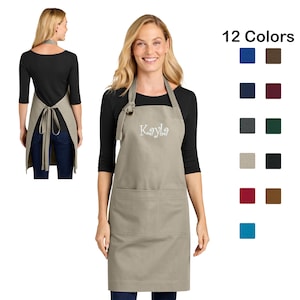 May include: A tan apron with the name "Kayla" embroidered in white. The apron has two front pockets and a neck loop. The image also shows a color chart with 12 color options.