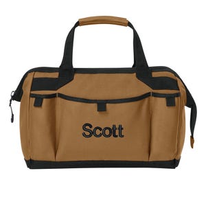 May include: A tan tool bag with black trim and black handles. The bag features multiple exterior pockets and the name Scott embroidered in black thread on the front center. The bag has a zippered top closure and a structured rectangular base.