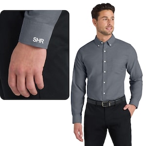 May include: A gray button-down shirt with a button-down collar and a chest pocket. The shirt is paired with black pants and a black belt. The cuff of the shirt has the initials "SHR" embroidered in white.