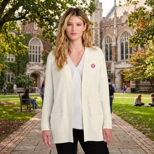 May include: A cream-colored, long-sleeved, open-front cardigan sweater featuring a ribbed knit texture, two front pockets, and a small embroidered pink daisy flower with a yellow center on the upper left chest area.