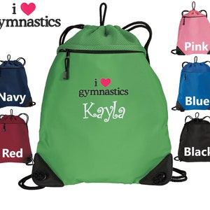 May include: Green drawstring bag with a zippered pocket and a mesh top. The bag has the text "i love gymnastics Kayla" embroidered on the front. The bag is shown with other drawstring bags in different colors: pink, blue, red, and black.