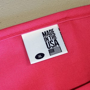 May include: Close-up of a bright pink garment with a white label. The label reads "MADE IN THE USA" with an American flag graphic and the size "XL". The fabric appears to be a smooth, woven material.