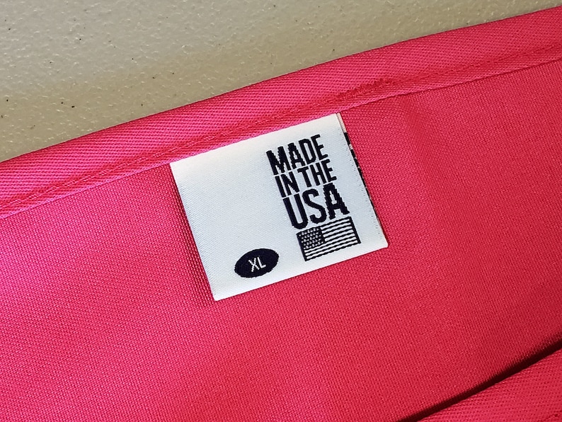 May include: Close-up of a bright pink garment with a white label. The label reads "MADE IN THE USA" with an American flag graphic and the size "XL". The fabric appears to be a smooth, woven material.