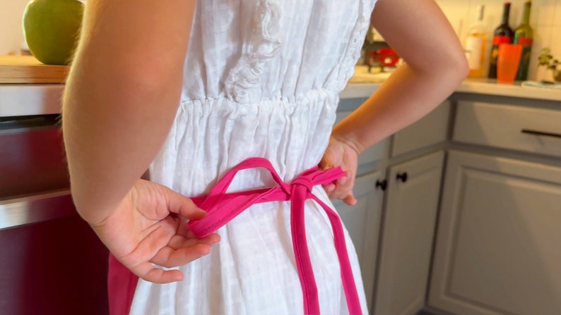 May include: A white dress with a pink apron tied in a bow. The apron's bright pink color contrasts with the white dress. The image is taken from behind, showing the apron being tied.