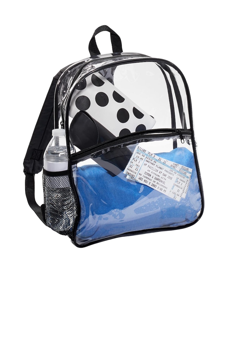 May include: A clear plastic backpack with black straps and a mesh side pocket. The backpack is filled with a blue towel, a water bottle, a black and white polka dot pouch, and a concert ticket for Simon & Garfunkel.
