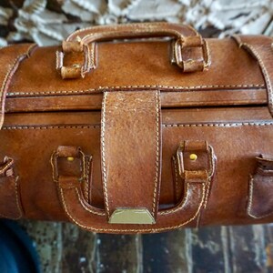 Puede incluir: Primer plano de una maleta vintage de cuero marr&oacute;n con asa y correas. La maleta tiene forma rectangular con costuras visibles y herrajes color lat&oacute;n. El cuero muestra signos de desgaste.