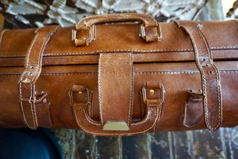 Puede incluir: Primer plano de una maleta vintage de cuero marr&oacute;n con asa y correas. La maleta tiene forma rectangular con costuras visibles y herrajes color lat&oacute;n. El cuero muestra signos de desgaste.