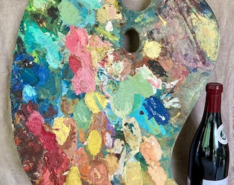 May include: A wooden artist's palette covered in a variety of dried paint colors, including pink, yellow, blue, and green. A bottle of red wine is next to the palette. The palette has a hole for the thumb and is approximately 12 inches long.