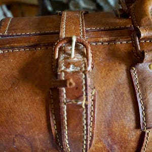 Puede incluir: Primer plano de una maleta vintage de cuero marr&oacute;n con cierre de hebilla. La maleta presenta costuras visibles y una hebilla de lat&oacute;n. El cuero tiene un aspecto desgastado, lo que sugiere edad y uso. La est&eacute;tica general es cl&aacute;sica y atemporal.