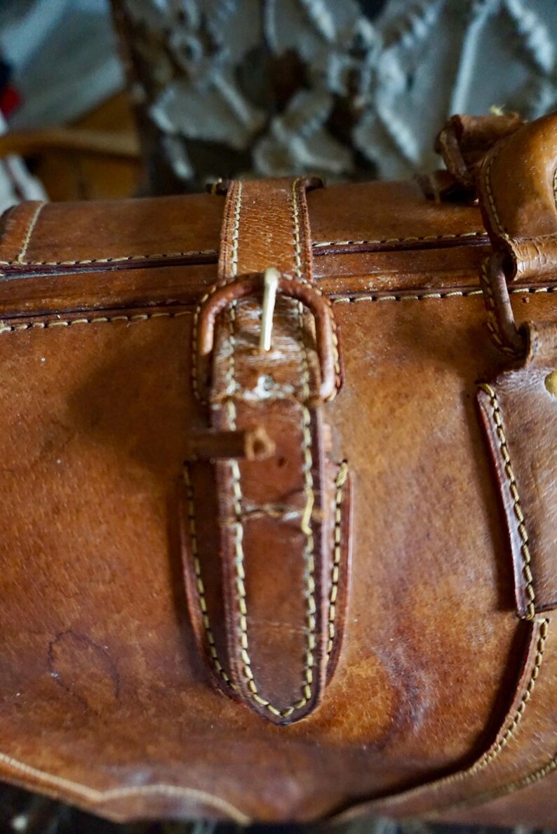 Puede incluir: Primer plano de una maleta vintage de cuero marr&oacute;n con cierre de hebilla. La maleta presenta costuras visibles y una hebilla de lat&oacute;n. El cuero tiene un aspecto desgastado, lo que sugiere edad y uso. La est&eacute;tica general es cl&aacute;sica y atemporal.
