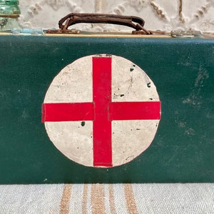 May include: A vintage green first aid kit, featuring a white circle with a red cross emblem. It has a brown handle and metal clasps. The aged appearance suggests it is an antique medical supply.