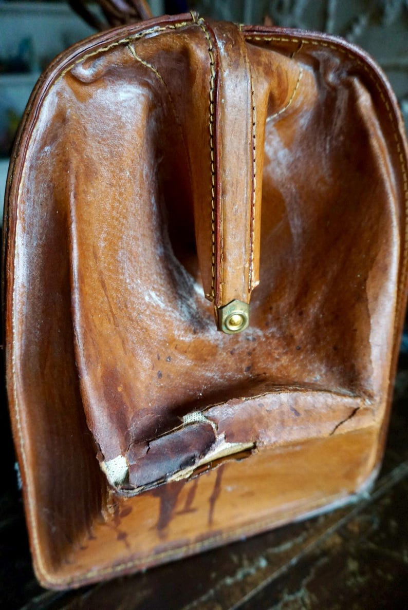 Puede incluir: Maleta de cuero marr&oacute;n vintage con asa y herrajes de lat&oacute;n. El equipaje usado muestra signos de edad, con desgaste visible en la superficie de cuero. El asa est&aacute; cosida y remachada.