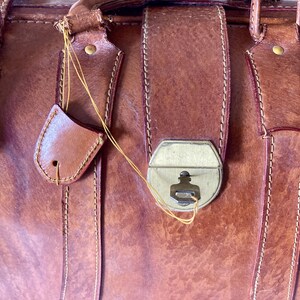 Puede incluir: Primer plano de un bolso de cuero marr&oacute;n vintage con un cierre de metal y correas de cuero. El bolso tiene un aspecto desgastado y texturizado con costuras visibles y una peque&ntilde;a etiqueta de cuero sujeta con una cuerda.